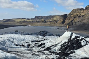 A hiker explores the Solheimajokull glacier with mountains and a small lagoon in the background.