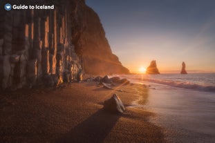 The sun peaking over the horizon over Reynifjara Black Sand Beach. Hexagonal basalt rock columns can be seen in the foreground to the left.