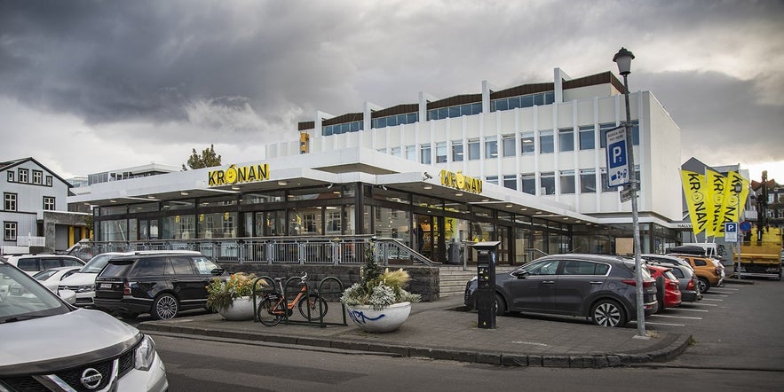 Krónan supermarket in Reykjavik with signage, parked cars, and yellow flags on a cloudy day.