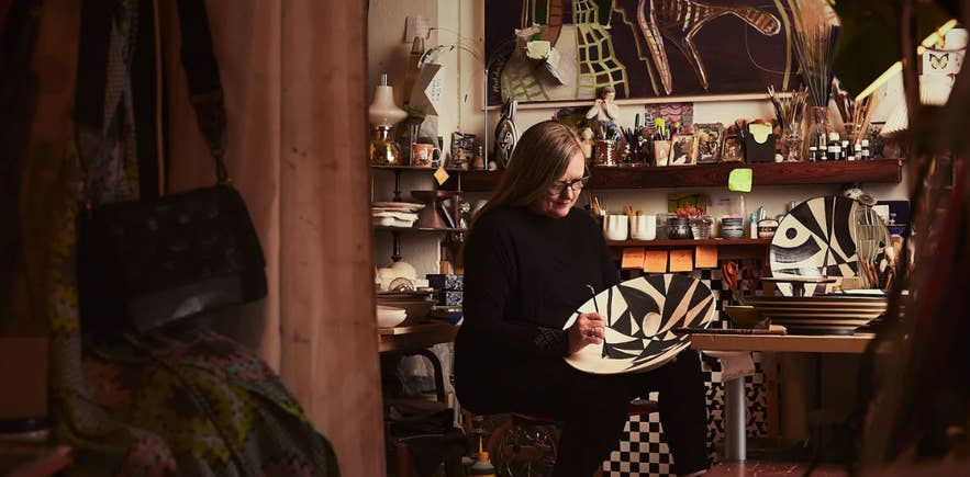 Inga Elín working in her Reykjavik art studio, hand-painting a large ceramic plate with bold black-and-white patterns, surrounded by colorful artwork, brushes, and pottery pieces at her namesake gallery.