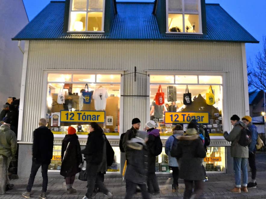 People gathered outside the iconic 12 Tónar record store in Reykjavik, Iceland, during evening hours, with music merchandise, vinyl, and CDs visible through large lit windows in a charming two-story house.