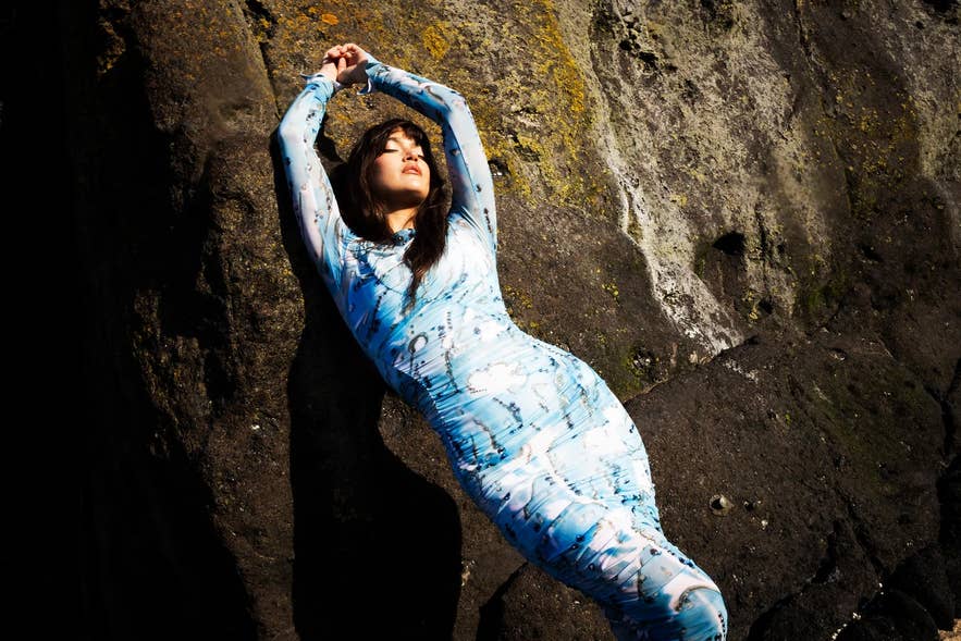 Model in figurbetontem, himmelblau bedrucktem Kleid der isländischen Modemarke Yeoman, posiert vor einer rauen Felswand im natürlichen Sonnenlicht.