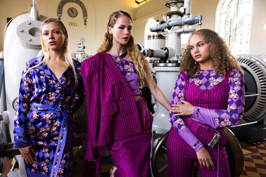 Modeller iført markante og farverige kreationer af den islandske designer Sif Benedicta, med lilla mønstre, nålestriber og sammensatte sæt, poserer i et interiør i industriel stil med vintage-maskiner.