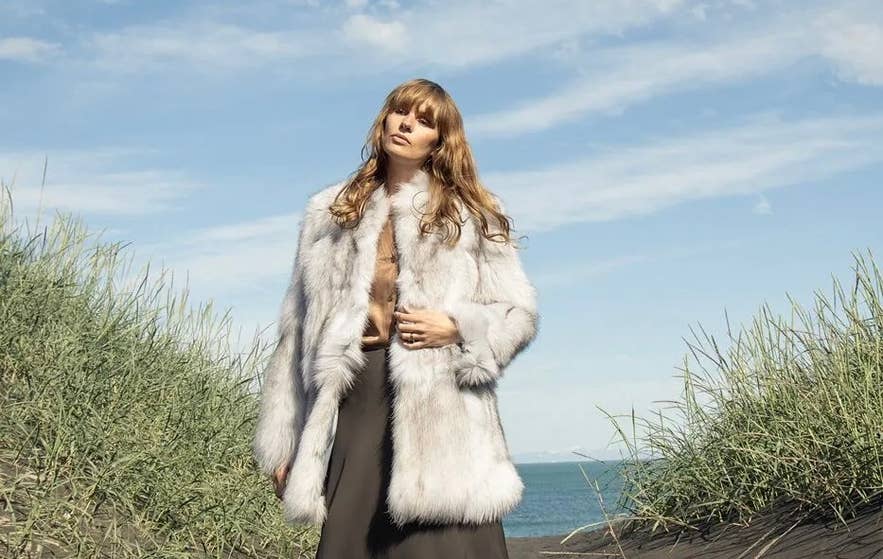En model iført en luksuriøs hvid pels fra Feldur Workshop, der poserer på en sort sandstrand i Island med højt grønt græs og en klarblå himmel i baggrunden.