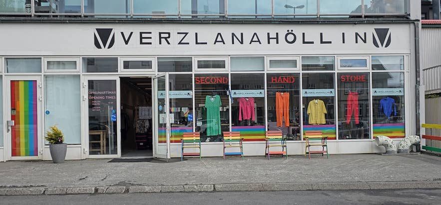 Udsiden af genbrugsbutikken Verzlanahollin i Reykjavik, Island, med tøjudstillinger i regnbuefarver, dekorationer med Pride-tema og en indbydende butiksfacade med farverige bænke og skilte.