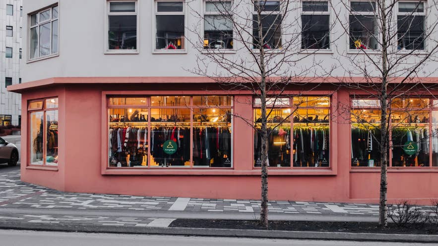 Buitenkant van Hringekjan kringloopwinkel in Reykjavik, IJsland, met warme verlichting, grote ramen gevuld met tweedehands kleding en een opvallende koraalroze gevel op een hoekpand.