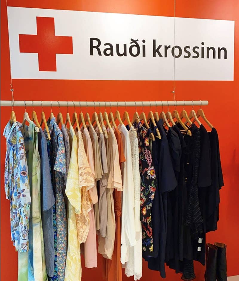 Et farverigt stativ med brugt tøj udstillet i Røde Kors-butikken (Raudi krossinn) i Reykjavik, Island, med den ikoniske røde kors-logo som baggrund på en knaldrød væg.