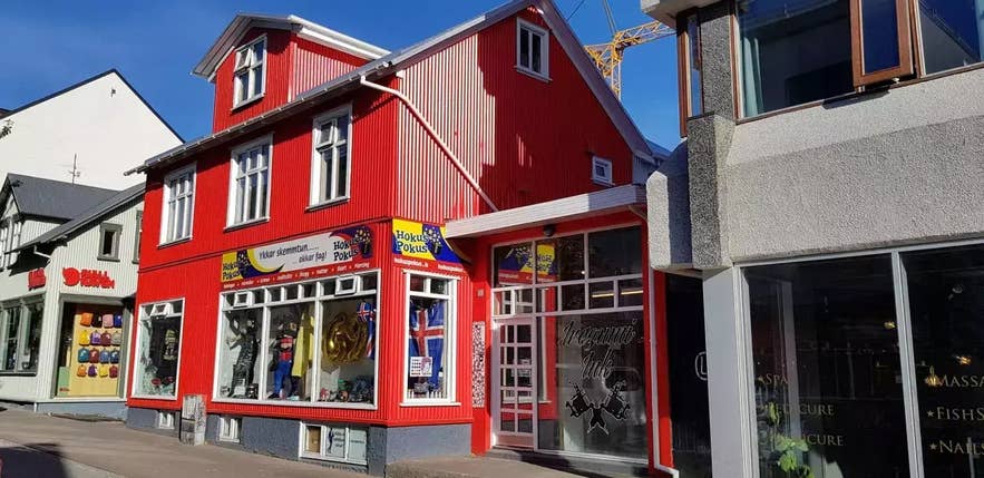 Udsiden af Hókus Pókus, en butik med magiske genstande og kostumer i Reykjavik, Island, der har til huse i en knaldrød, traditionel islandsk bygning med farvestrålende skilte og udstillingsvinduer fyldt med teatermasker, rekvisitter og souvenirs.