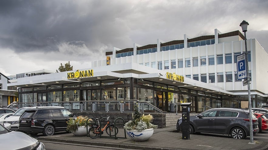 Exterior of Krónan grocery store in Reykjavik, Iceland, with modern architecture, large windows, and a street view featuring parked cars, bicycles, and dramatic cloudy skies.