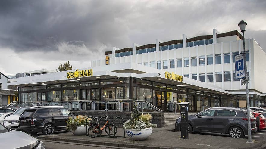 Udsiden af Krónan-købmandsbutikken i Reykjavik, Island, med moderne arkitektur, store vinduer og en udsigt over gaden med parkerede biler, cykler og en dramatisk overskyet himmel.