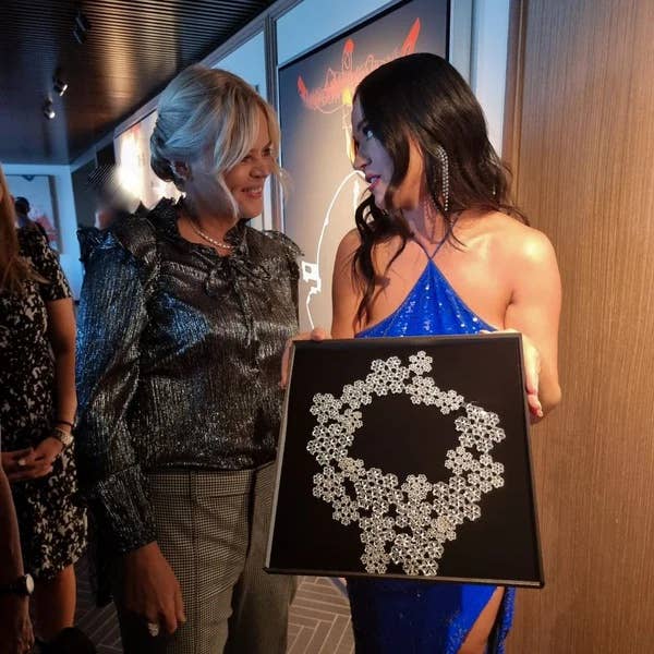 Katy Perry hält eine gerahmte isländische Halskette von Aurum Jewelry in den Händen, trägt ein glitzerndes blaues Kleid und unterhält sich mit einer Vertreterin bei einer Veranstaltung.