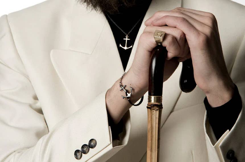 Close-up of a man in a white suit holding a cane, wearing an anchor necklace and matching anchor bracelet from Icelandic jewelry brand Orrifinn, along with a gold signet ring.