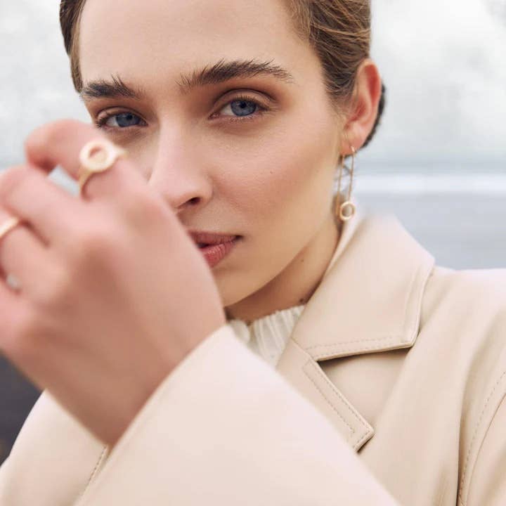 Close-up of a woman wearing minimalist gold jewelry from the Icelandic brand Mjöll, including a circular statement ring and matching dangling earrings, styled with a beige coat in soft natural light.
