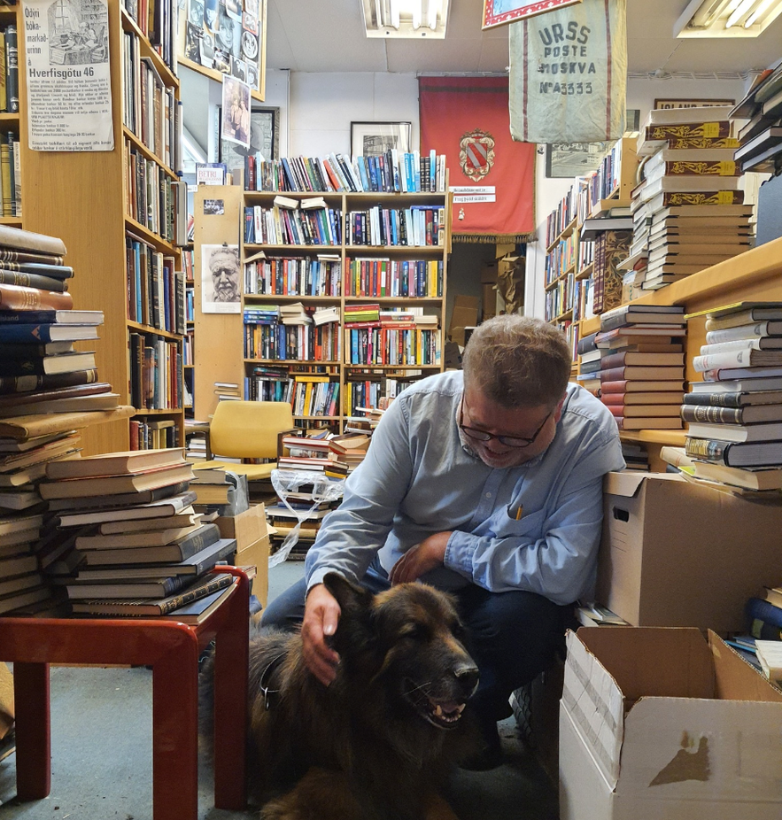 Man petting a German Shepherd inside Bókin, a secondhand bookstore in Reykjavik, Iceland, surrounded by towering stacks of books and a cozy, cluttered literary atmosphere.