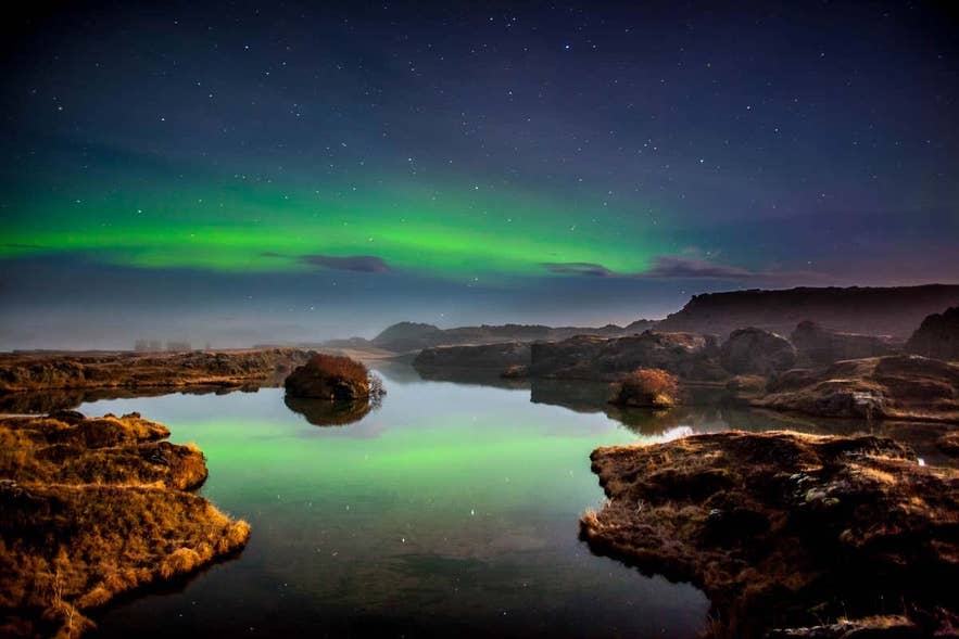 Northern lights near Akureyri with calm lake reflections, rocky landscape, and starry night sky in Iceland