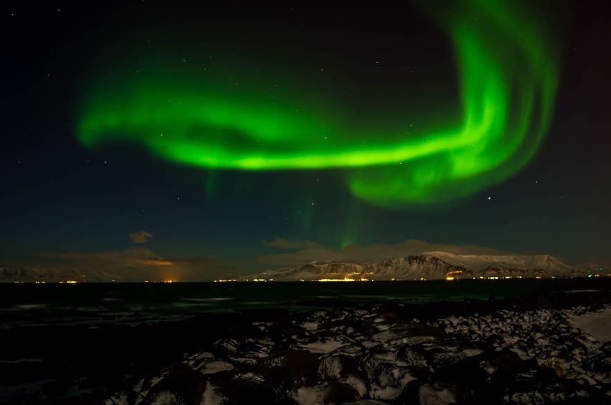 Aurores boréales au-dessus de la côte de Reykjavik, avec un premier plan enneigé et les lumières de la ville sous un ciel nocturne vert.