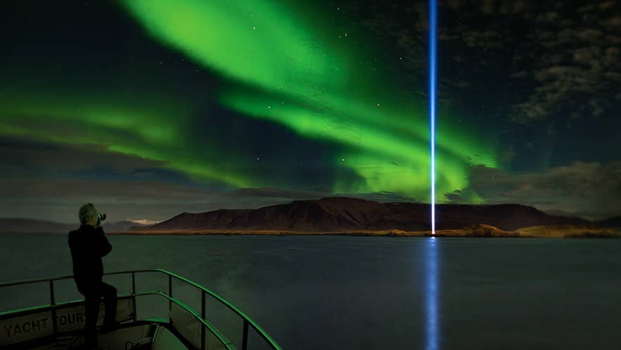 Northern lights over Reykjavik sea with Imagine Peace Tower beam, aurora reflecting on water and person viewing from boat