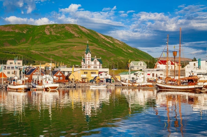 Colorful houses and whale-watching boats fill H&uacute;sav&iacute;k harbor, backed by green hills and bright summer skies.