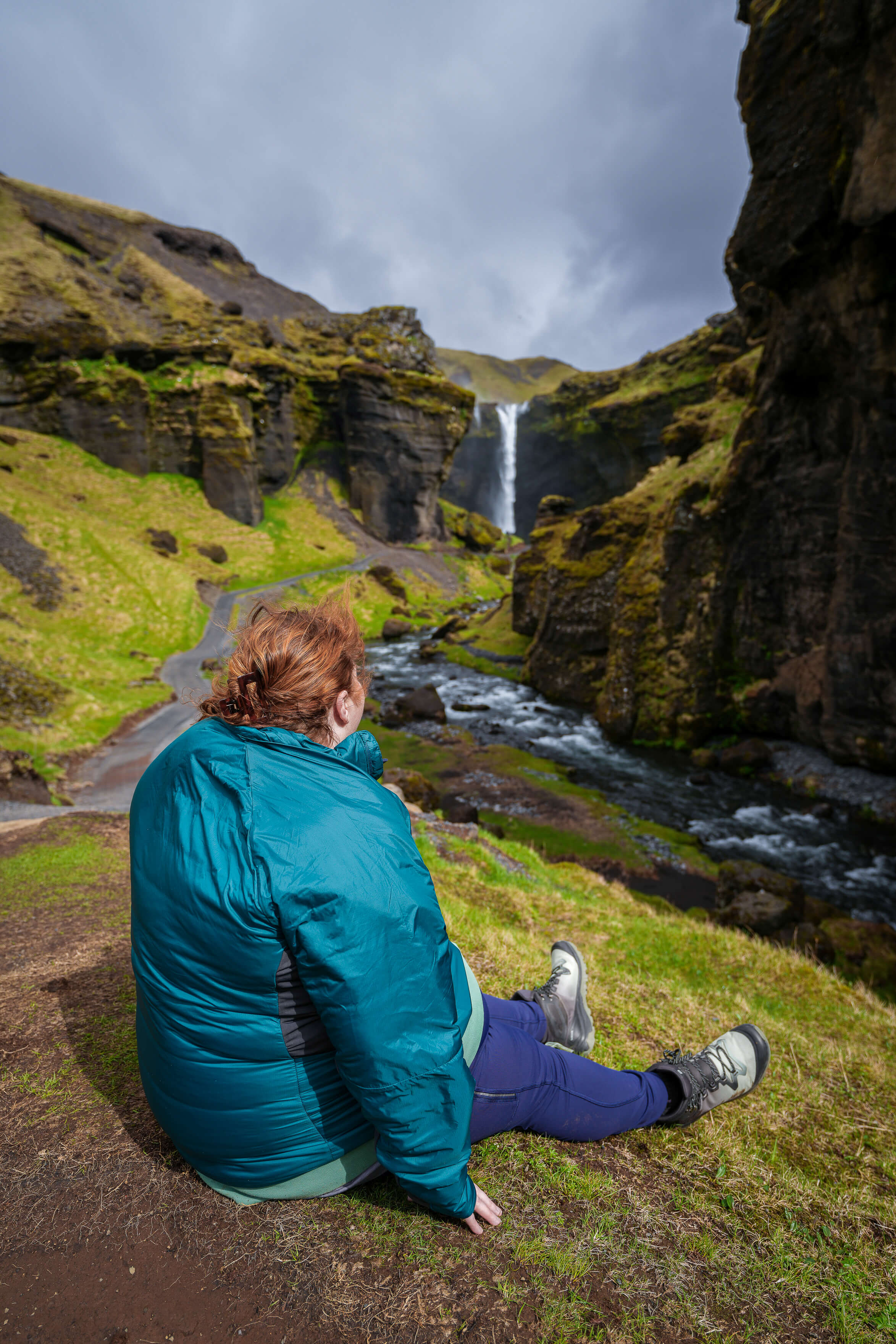 A woman sits on the ground while gazing out at Kvernufoss  Waterfall in the distance.