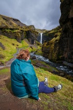 A woman sits on the ground while gazing out at Kvernufoss  Waterfall in the distance.
