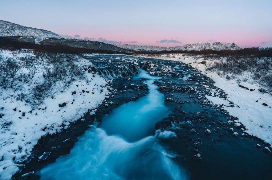 冬のブルアゥルフォス滝。雪に覆われた岸辺と凍った景色、薄明かりの空。