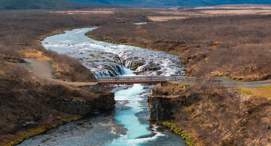 Aerial view of Bruarfoss waterfall and the footbridge crossing its brilliant blue stream, surrounded by barren early spring landscape.