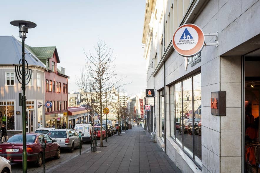Вид на улицу с HI Loft Hostel в Рейкьявике, Исландия, с вывеской Hostelling International, припаркованными автомобилями и магазинами на улице Лаугавегур.