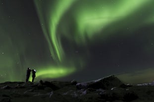 Two people stand on top of a small hill admiring the northern lights.