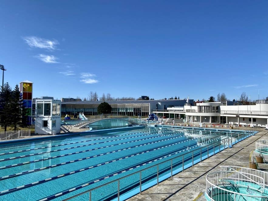 Piscina Laugardalslaug a Reykjavik, Islanda, con piscine geotermiche all'aperto, scivoli d'acqua e corsie per il nuoto sotto un cielo azzurro.