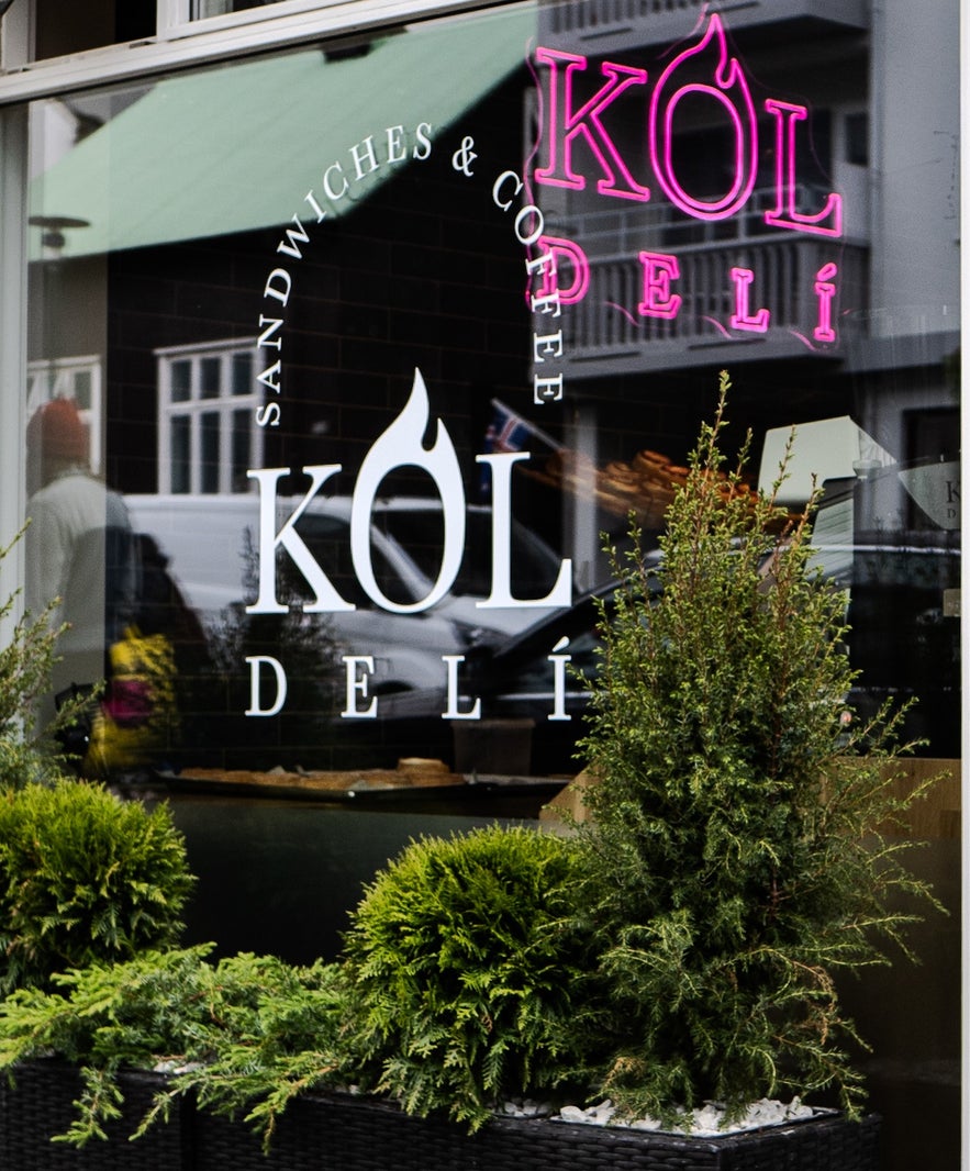 Exterior of Kol Deli in Reykjavik with logo on window and green plants in front.