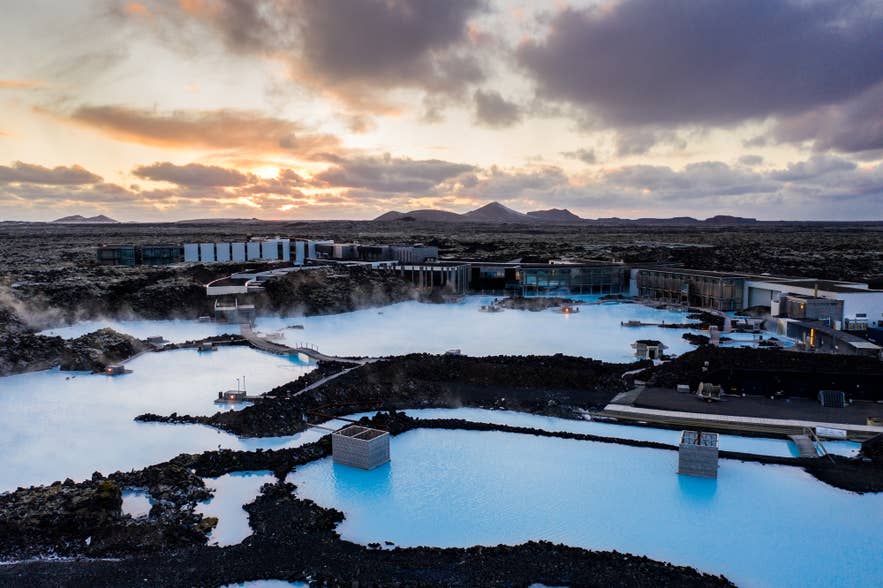 Vue aérienne du spa géothermique Blue Lagoon en Islande au coucher du soleil, avec ses eaux bleu laiteux fumantes entourées de champs de lave volcanique et d’installations de luxe.