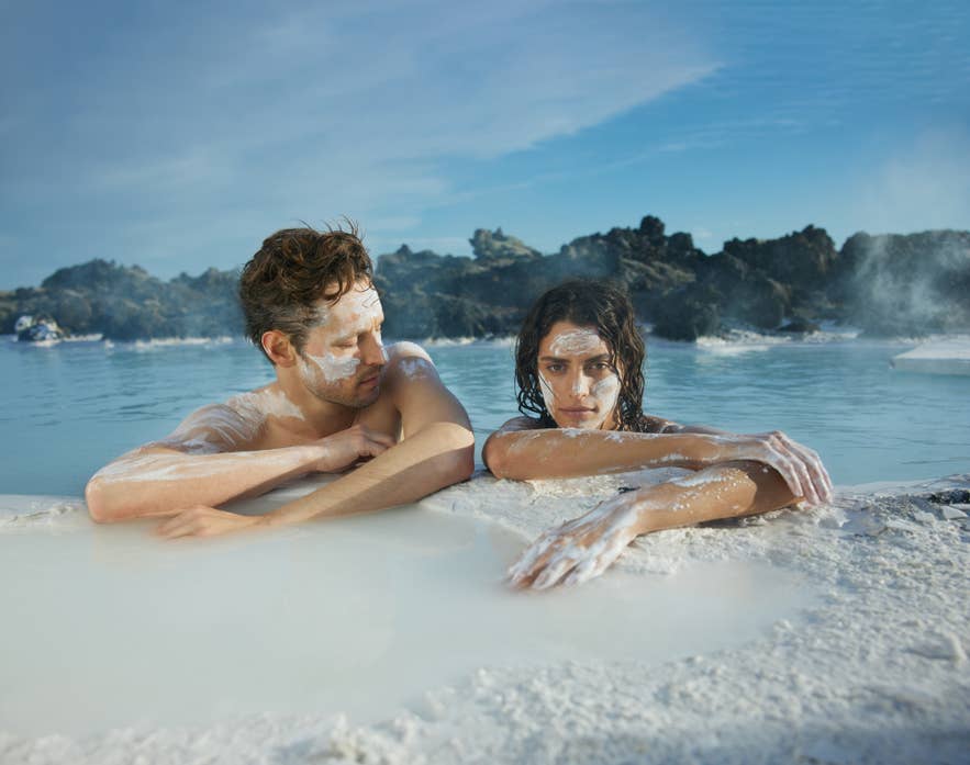 Pareja relaj&aacute;ndose en el balneario geot&eacute;rmico de la Laguna Azul en Islandia, con mascarillas blancas de s&iacute;lice en la cara y agua azul mineral rodeada de rocas volc&aacute;nicas.