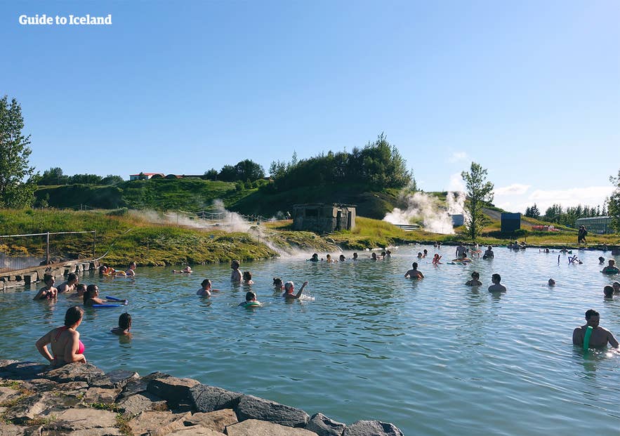 Locals and tourists bathing in the Secret Lagoon geothermal hot spring in Iceland, surrounded by lush green hills and steam vents on a sunny summer day.