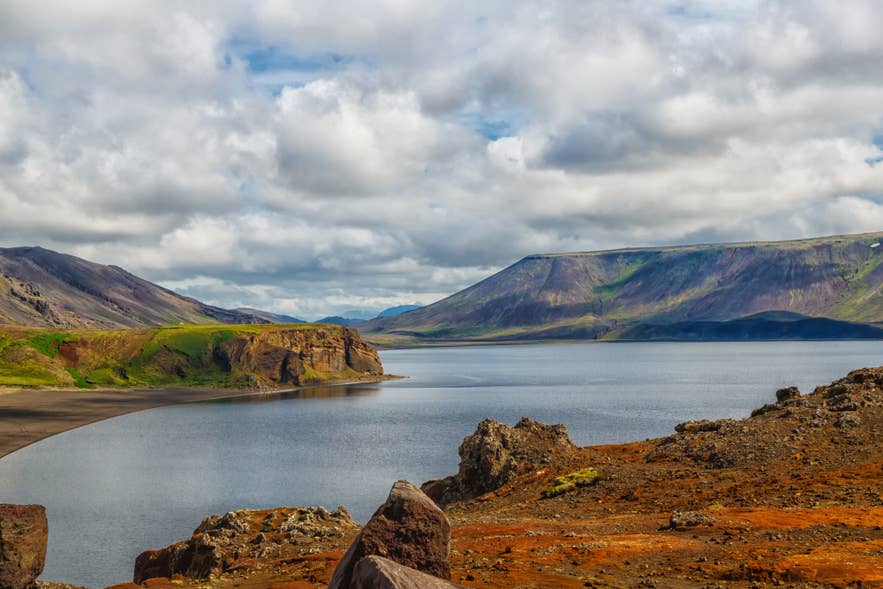 Colorful volcanic landscape with red lava rocks and rugged mountains surrounding Kleifarvatn Lake on the Reykjanes Peninsula in Iceland, under a dramatic cloudy sky.