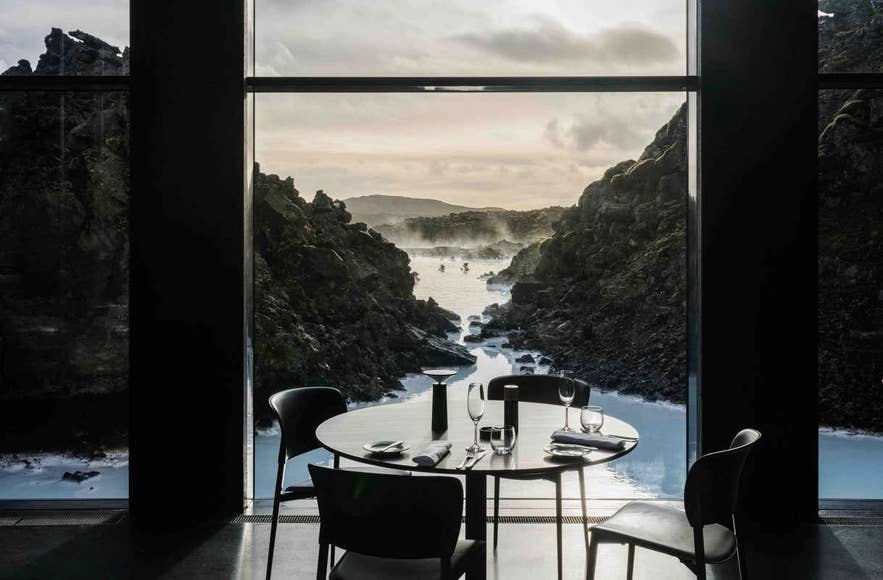 Mesa elegante con copas de vino con vistas a las aguas geot&eacute;rmicas y campos de lava volc&aacute;nica de la Laguna Azul en Islandia, vista a trav&eacute;s de una gran ventana panor&aacute;mica en el Lava Restaurant.