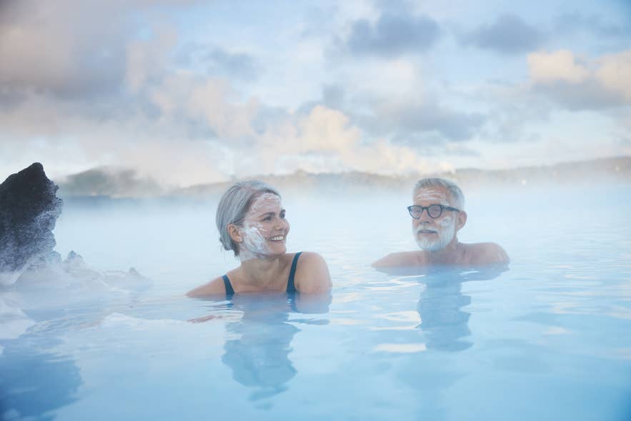 Ouder echtpaar dat glimlachend geniet van het warme water van de Blue Lagoon in IJsland, beiden met maskers van kiezelmodder op hun gezicht onder een bewolkte hemel.