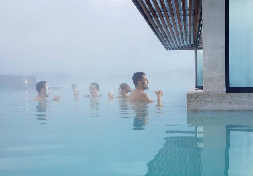 Group of people enjoying drinks and silica masks in the steaming waters of the Blue Lagoon geothermal spa in Iceland, beside a modern glass-walled structure.