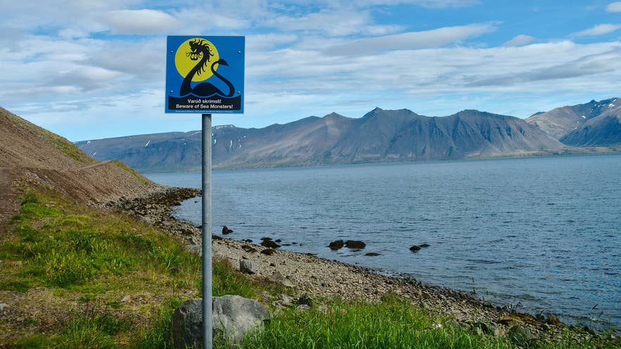 Warning sign reading “Beware of Sea Monsters” by the shoreline of Arnarfjörður fjord in the Westfjords of Iceland, with mountains and calm water in the background. Warning sign reading “Beware of Sea Monsters” by the shoreline of Arnarfjörður fjord in the Westfjords of Iceland, with mountains and calm water in the background.