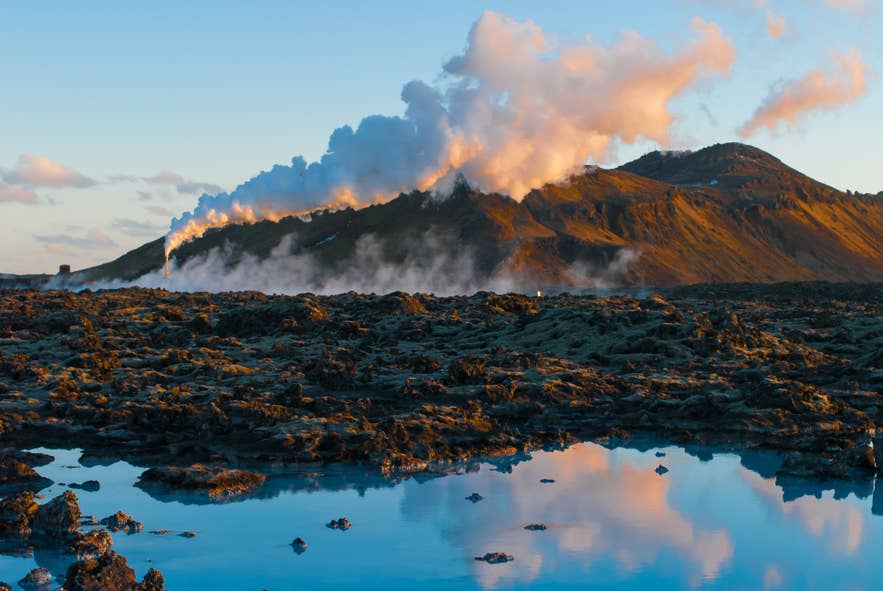 Geothermal steam rising from the Svartsengi power plant in Iceland at sunset, with dramatic clouds and volcanic moss-covered lava fields in the foreground.