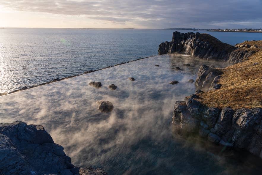 Luchtfoto van het geothermische overloopzwembad van Sky Lagoon in IJsland, met opstijgende stoom tegen de achtergrond van de Atlantische Oceaan bij zonsondergang, waar natuurlijke kliffen samengaan met modern spa-ontwerp.