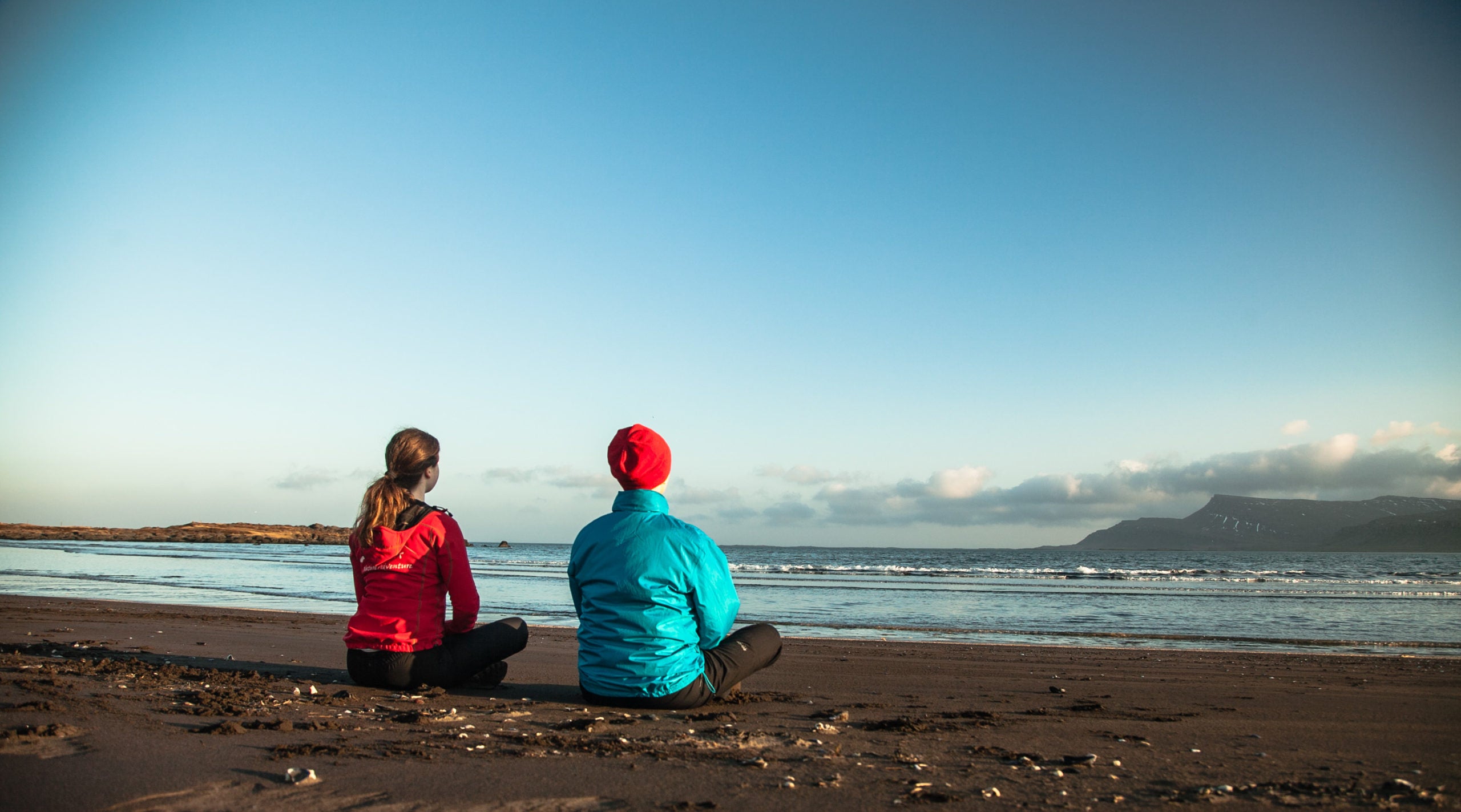 Two people sit cross-legged on a black sand beach during a yoga tour in East Iceland.