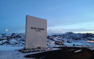 A tall white sign reading 'Blue Lagoon Iceland' stands among snow-covered lava rocks under a dusky winter sky.