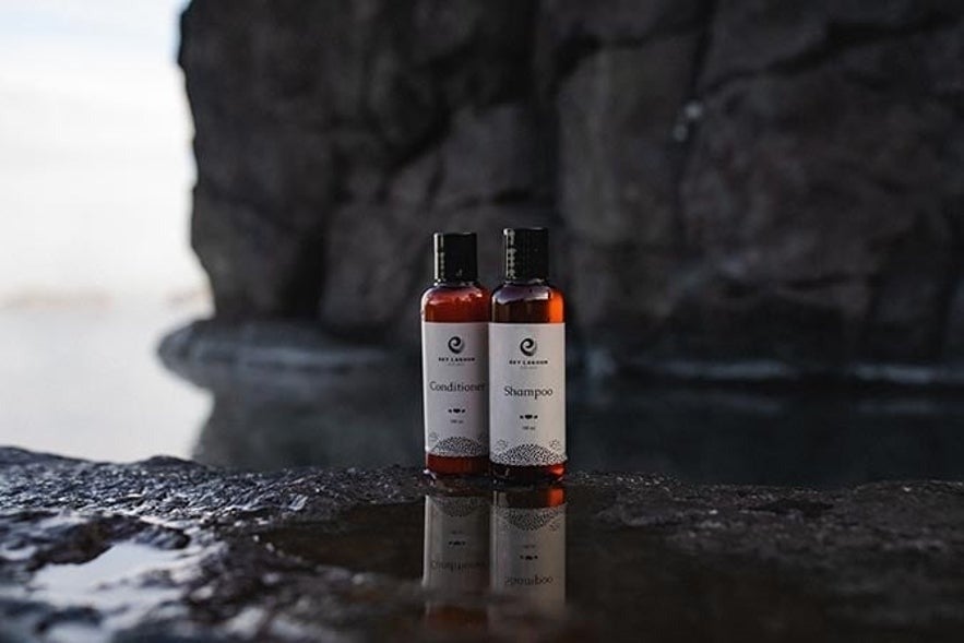Sky Lagoon-branded shampoo and conditioner bottles placed on wet volcanic rock with geothermal steam and dramatic cliffside in the background, capturing the natural Icelandic spa experience.