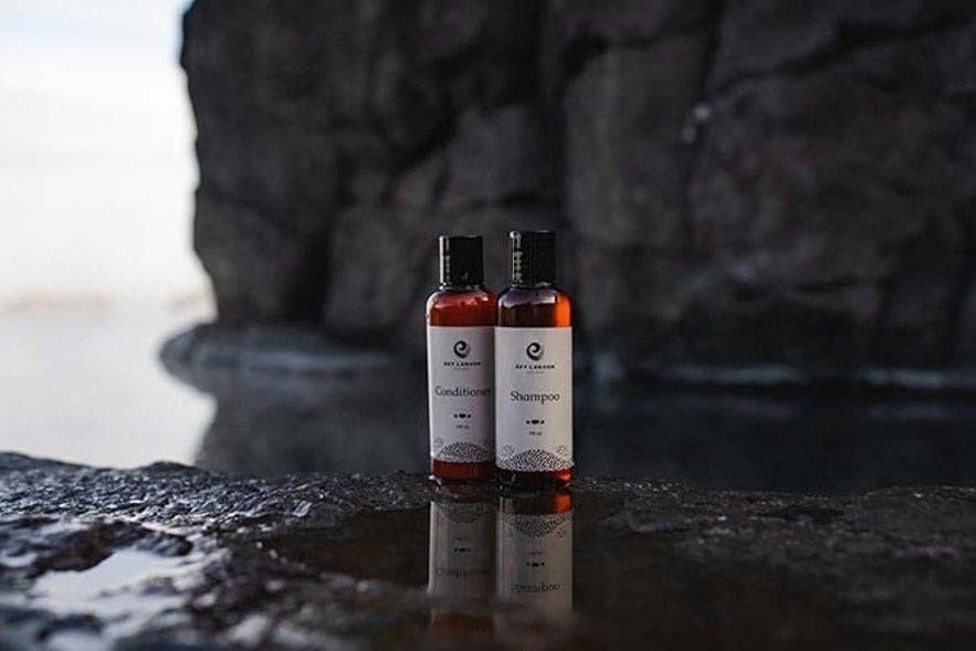 Flasker med shampoo og balsam fra mærket Sky Lagoon placeret på våd vulkansk sten med geotermisk damp og en dramatisk klippekant i baggrunden, hvilket indfanger den naturlige islandske spa-oplevelse.