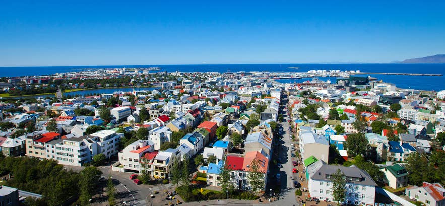 Panoramaudsigt over Reykjaviks centrum i Island med farverige hustage, bybilledet ved kysten og Nordatlanten under en klar blå himmel.