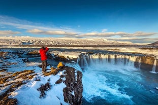 A man wearing a red jacket takes a picture of the beautiful Godafoss waterfall.