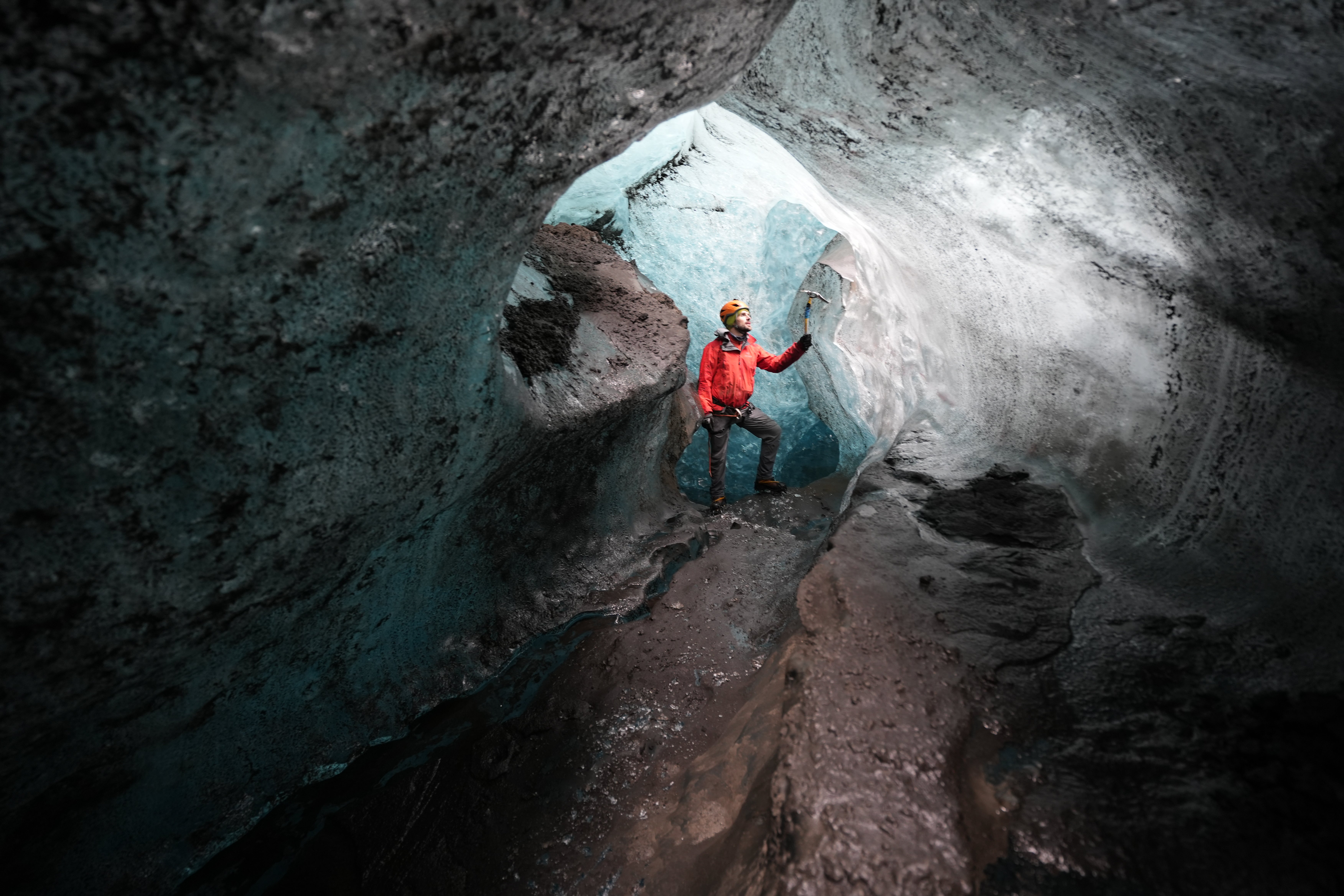 A man wearing an orange jacket stands inside a beautiful Ice Cave in Iceland.