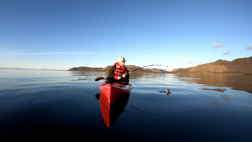 Man fishing from a red kayak on calm waters in Iceland in June, surrounded by mountains under clear skies during a peaceful summer evening.