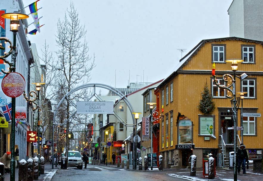 Via dello shopping Laugavegur a Reykjavik, Islanda, in inverno, con edifici colorati, negozi, lampioni, decorazioni natalizie, neve e passanti sotto un cielo nuvoloso.