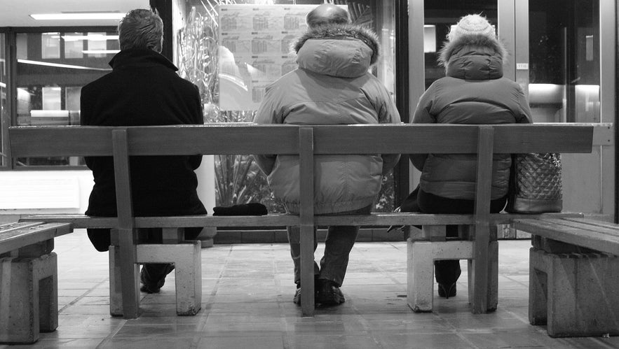 Black-and-white photo of three people waiting for a bus at Hlemmur terminal in Reykjavík, Iceland, wearing winter jackets, sitting on a wooden bench indoors.